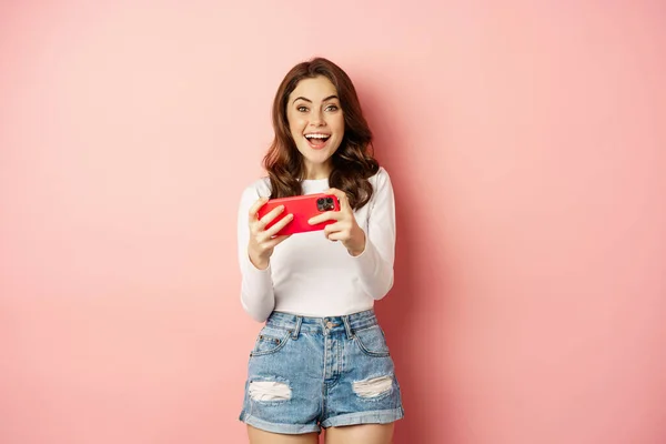Menina bonita feliz rindo e sorrindo, assistindo vídeo, jogando jogos no telefone celular, smartphone horizontal, de pé sobre fundo rosa — Fotografia de Stock