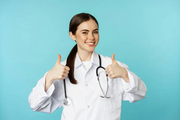 Happy woman doctor, medical worker in white coat, showing thumbs up in approval, like something, praise, standing over blue background — Fotografia de Stock