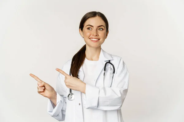 Porträt einer lächelnden jungen Ärztin, medizinischen Fachangestellten, die mit dem Finger nach links zeigt, Klinik-Promo, Logo oder Banner zeigt, auf weißem Hintergrund stehend — Stockfoto