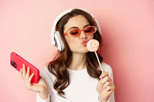 Close up portrait of stylish glamour girl, beautiful woman in headphones, listening music with smartphone, wearing sunglasses and licking lolipop — Zdjęcie stockowe