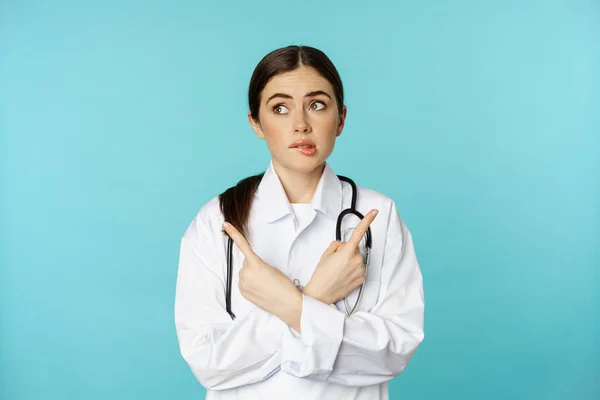 Portrait of indecisive, confused female doctor, pharmacy worker pointing sideways and biting lip clueless, dont know, standing over blue background — Zdjęcie stockowe