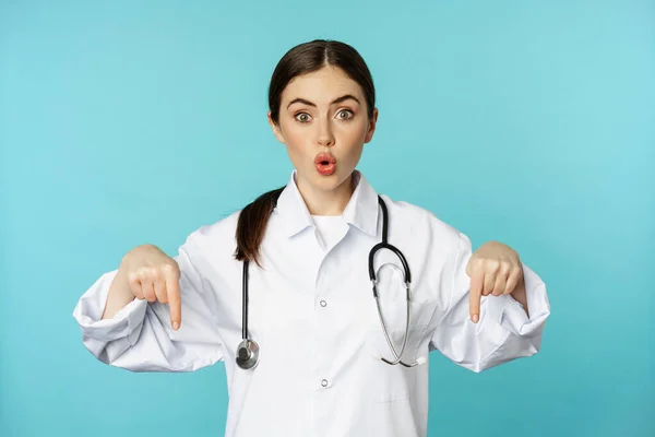 Enthusiastic healthcare worker, young woman doctor in white coat, pointing fingers down and smiling, showing pharmacy advertisement, medical promo, blue background — Stok fotoğraf
