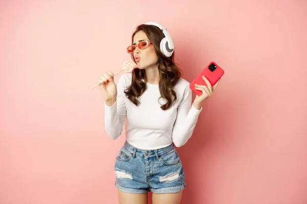 Summer girl laughing, eathing lolipop and listening music in headphones, dancing with smartphone against pink background — Zdjęcie stockowe