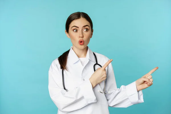 Portrait of surprised medical worker, woman doctor pointing fingers right and say wow amazed, reacting interested, standing over torquoise background — ストック写真