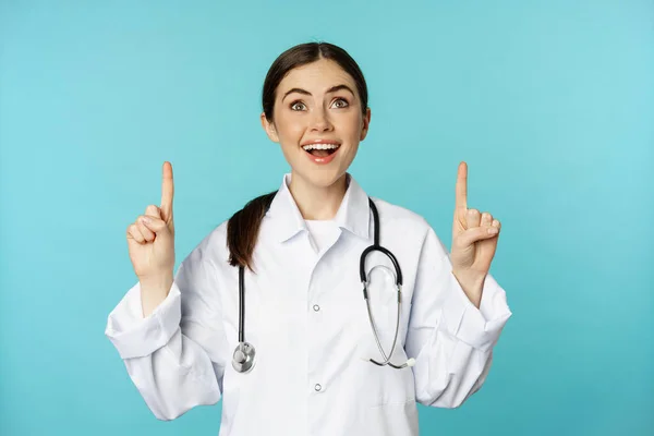 Enthusiastic medical worker, young woman doctor in white coat, stethoscope, showing advertisement, pointing fingers up, standing over torquoise background — Stok fotoğraf