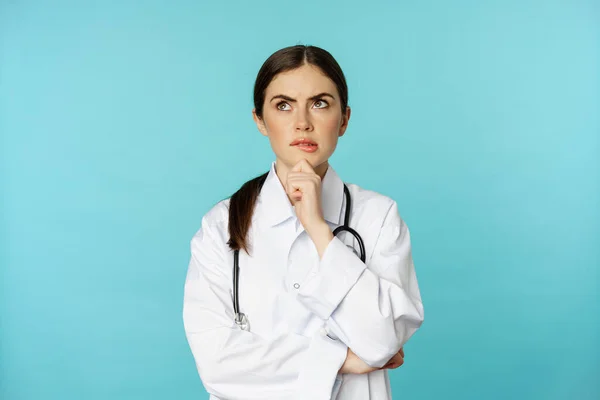 Mladá doktorka, nemocniční pracovnice v bílém plášti, přemýšlivá a zamyšlená, hledající řešení, stojící nad pozadím toquoise — Stock fotografie