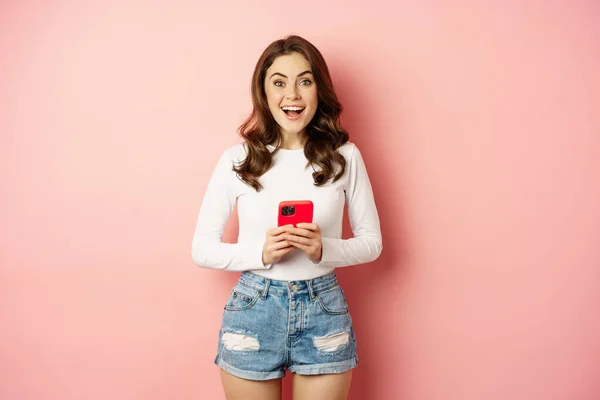 Conceito de compras online e celular. Garota atraente animado segurando telefone celular e sorrindo, de pé sobre fundo rosa — Fotografia de Stock