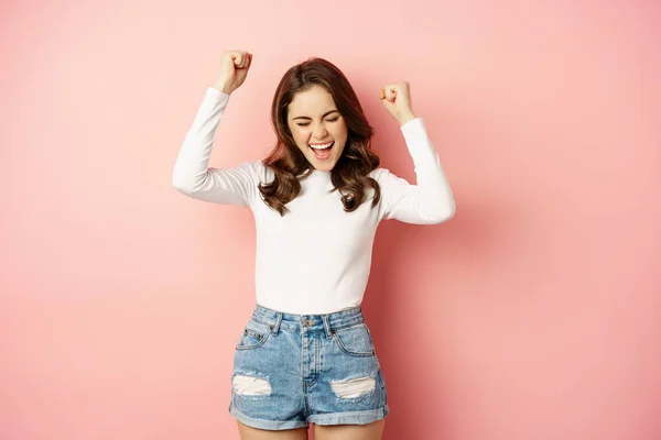 She is a winner. Enthusiastic brunette girl celebrating, jumping from happiness and shouting joyful, achieve goal success, standing over pink background — Fotografia de Stock
