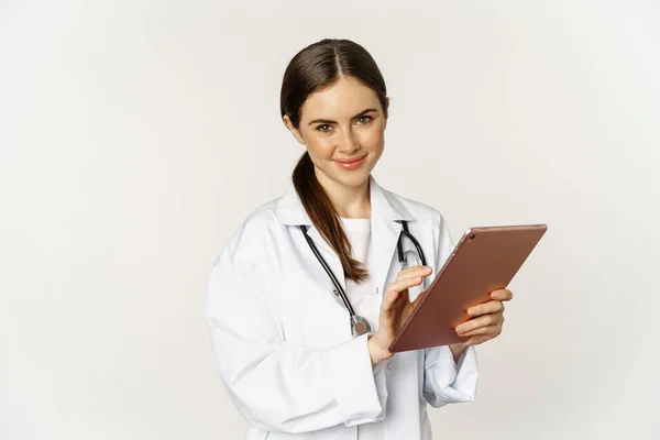 Doctor in white coat using digital tablet, reading medical data on gadget, working in hospital, standing over white background — Foto de Stock