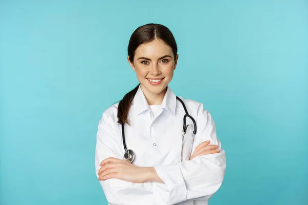 Medical staff and doctors concept. Young smiling female doctor, healthcare worker in white coat and stethoscope, looking confident, waiting patients, blue background — ストック写真