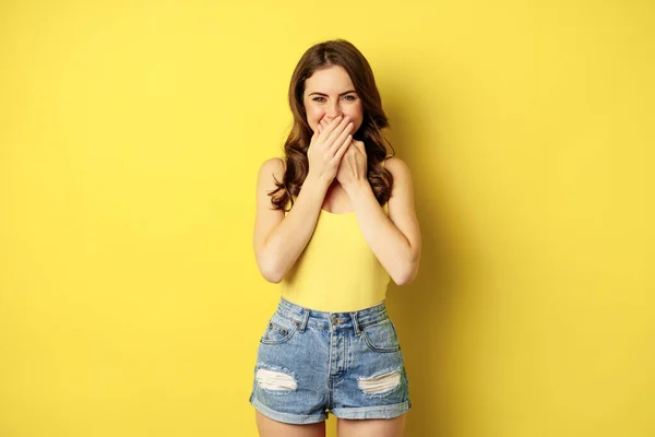 Beautiful coquettish woman giggle, laughing and covering mouth with hands, smiling with eyes, standing over yellow background — Stok fotoğraf