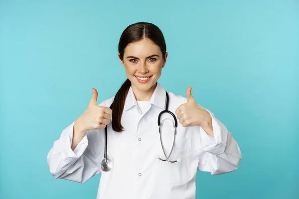 Happy woman doctor, medical worker in white coat, showing thumbs up in approval, like something, praise, standing over blue background — ストック写真
