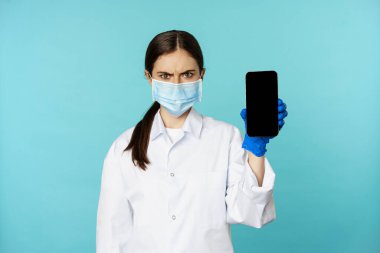 Angry and confused woman doctor in face mask, gloves, showing smartphone app, mobile screen, frowning upset, standing over blue background