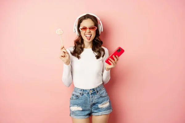Mujer morena con estilo bailando en auriculares con teléfono móvil, escuchando música en auriculares, lamiendo piruleta, de pie sobre fondo rosa — Foto de Stock
