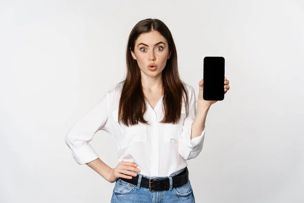 Enthusiastic corporate woman demonstrating website, mobile phone screen, showing application and saying wow, standing over white background — Fotografia de Stock