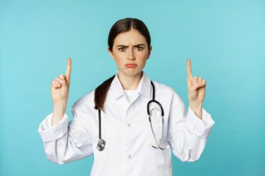 Sad and disappointed healthcare worker, doctor pointing fingers up, frowning upset, showing bad news, standing over torquoise background