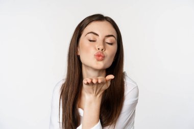 Feminine woman sending air kiss at camera, coquettish flirty pose, kissing, standing over white background