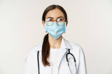 Portrait of medical worker, female physician in face mask from covid during pandemic, smiling and looking enthusiastic, standing over white background