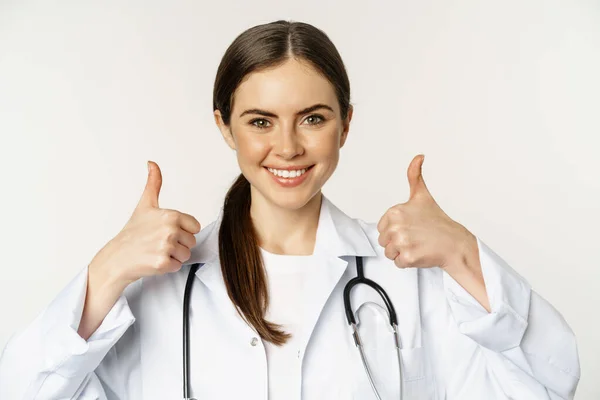 Porträt einer lächelnden schönen Ärztin im weißen Mantel, die die Daumen nach oben zeigt, vor weißem Hintergrund stehend — Stockfoto