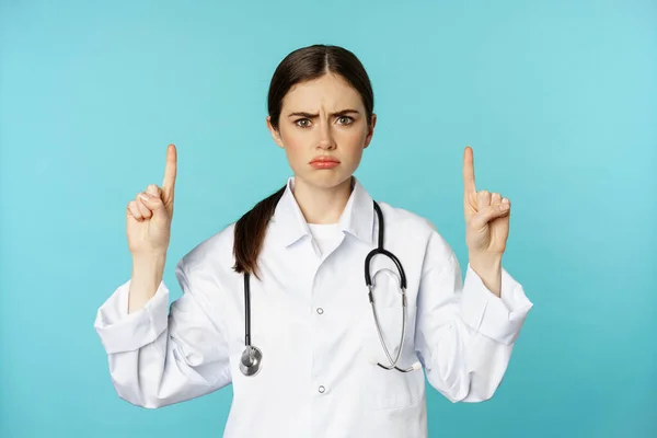 Sad and disappointed healthcare worker, doctor pointing fingers up, frowning upset, showing bad news, standing over torquoise background — Stok fotoğraf