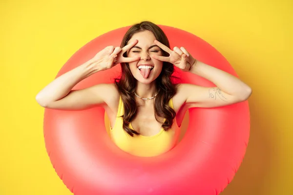 Portret atrakcyjnej nowoczesnej dziewczyny wewnątrz modnej różowej plaży pierścień pływacki, pokazując spokój v-sign i uśmiechnięty szczęśliwy, stojąc na żółtym tle — Zdjęcie stockowe