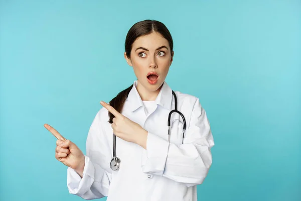 Surprised female doctor, physician with stethoscope, pointing and looking left with amazed, wow face, standing over torquoise background — Stok fotoğraf