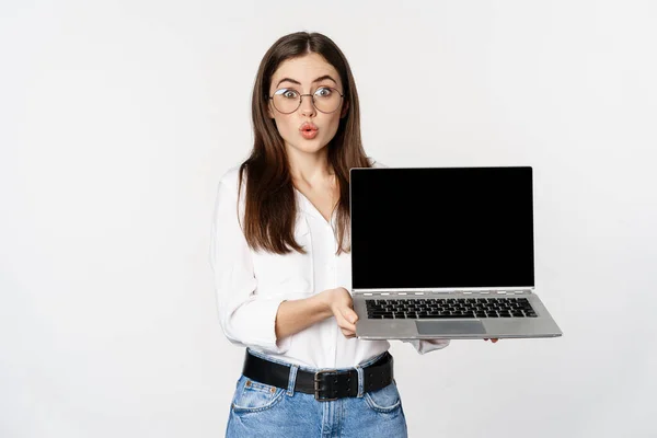 Портрет милой девушки в очках, студент показывает экран ноутбука с изумленным лицом, стоя на белом фоне — стоковое фото