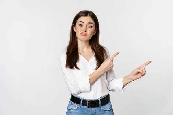 Sad and disappointed cute woman, gloomy girl pointing fingers at logo, complaining, sulking upset, standing in blouse over white background — 图库照片