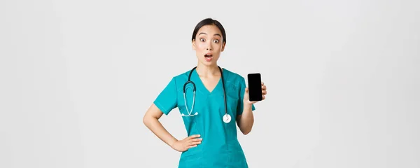 Covid-19, εργαζόμενοι στον τομέα της υγείας και online ιατρική έννοια. Ενθουσιασμένος και έκπληκτος ασιατική νοσοκόμα, γιατρός φαίνονται έκπληκτοι, ενώ δείχνει οθόνη του κινητού τηλεφώνου, internet app διαβούλευση, λευκό φόντο — Φωτογραφία Αρχείου