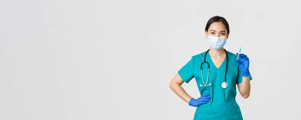 Covid-19, onemocnění koronavirem, koncept zdravotnických pracovníků. Usmívající se sebevědomá asijská ošetřovatelka v gumách a lékařské masce, držící stříkačku s vakcínou, očkovací injekce, bílé pozadí — Stock fotografie