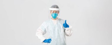 Covid-19, koronavirüs hastalığı, sağlık çalışanları konsepti. Kişisel koruma ekipmanları içinde ciddi görünümlü Asyalı kadın doktor, baş parmağını kaldırıyor, hastanın güvenliğini sağlıyor.