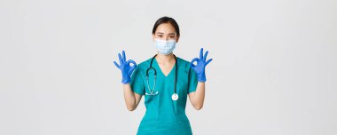 Covid-19, koronavirüs hastalığı, sağlık çalışanları konsepti. Gülümseyen profesyonel ve kendinden emin Asyalı doktor, tıbbi maske, eldiven ve önlük giyen doktor iyi niyet göstergesi, kaliteli hizmet garantisi