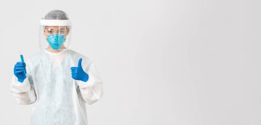 Covid-19, koronavirüs hastalığı, sağlık çalışanları konsepti. Ciddi ciddi Asyalı kadın doktor, kişisel koruma ekipmanlarında teknoloji laboratuarı çalışanı baş parmağını kaldır ve aşı ile test tüpünü tut.