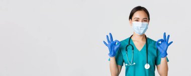 Covid-19, koronavirüs hastalığı, sağlık çalışanları konsepti. Kendinden emin gülümseyen Asyalı kadın doktor, tıbbi maske ve eldivenli hemşire, onaylı, beyaz arka planda iyi niyet göstergesi göster.