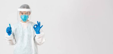 Covid-19, koronavirüs hastalığı, sağlık çalışanları konsepti. Ciddi bir Asyalı kadın doktor, kişisel koruyucu ekipman araştırmacısı iyi bir jest gösteriyor, elinde aşı ile test tüpü tutuyor.