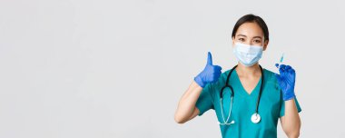 Covid-19, koronavirüs hastalığı, sağlık çalışanları konsepti. Gülümseyen güzel Asyalı tıp çalışanı, maskeli hemşire ve eldivenli başparmak kaldıran, aşılı şırıngayı tutan, ayakta duran beyaz arka plan