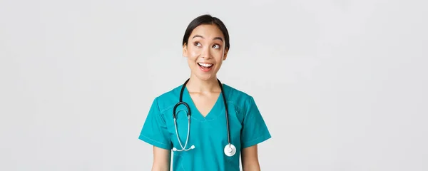 Sjukvårdspersonal, förebyggande av virus, karantänkampanjkoncept. Entusiastiska leende asiatisk läkare i buskar, sjuksköterska tittar övre vänstra hörnet med roat ansikte, stående vit bakgrund — Stockfoto
