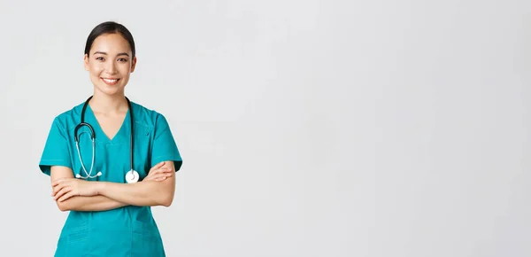 Covid-19, Gesundheitspersonal, Pandemiekonzept. Porträt einer selbstbewusst lächelnden, attraktiven asiatischen Krankenschwester im Peeling, mit Stethoskop, Kreuzarmbrust und Blick in die Kamera, weißer Hintergrund — Stockfoto