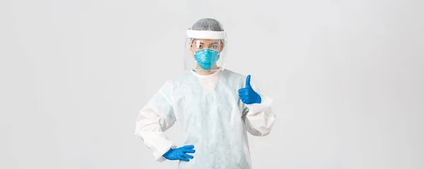 Covid-19, koronavírus betegség, egészségügyi dolgozók koncepciója. Súlyos kinézetű, professzionális ázsiai női orvos egyéni védőfelszerelésben, hüvelykujjával felfelé mutatva, biztosítja a beteg biztonságát — Stock Fotó