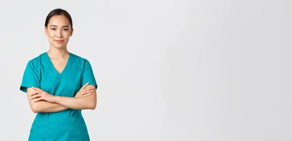 Covid-19, zdravotníci, pandemický koncept. Sebevědomě se usmívající asijská ošetřovatelka v křoví, stojící sebevědomě, se zkříženýma rukama. Profesionální lékař připraven na operaci, stojící bílé pozadí — Stock fotografie