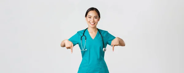 Covid-19, egészségügyi dolgozók, világjárvány. Mosolygó kellemes ázsiai női orvos, terapeuta vagy orvos köpenyben sztetoszkóppal, ujjal lefelé, show klinika banner — Stock Fotó