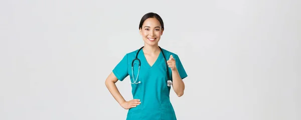 Covid-19, sjukvårdspersonal, pandemikoncept. Självsäker leende asiatisk professionell läkare, sjuksköterska i skrubbar pekande finger mot kameran, inbjudande för kontroll eller vaccination, ge rekommendation — Stockfoto
