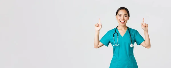 Covid-19, trabajadores de la salud, concepto pandémico. Sonriente enfermera bonita alegre, doctora o pasante en matorrales apuntando con los dedos hacia arriba, mostrando pancarta, hacer anuncio, demostrar publicidad — Foto de Stock