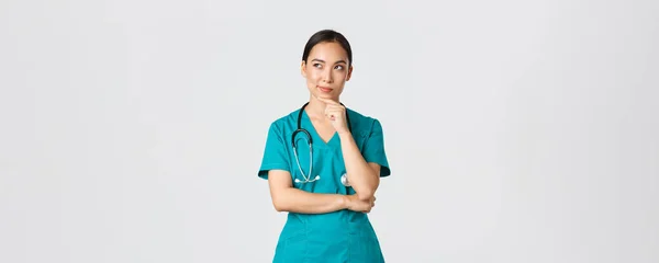 Ковід-19, медичні працівники, пандемія. Розумна азіатська медсестра в скрабах дивиться в очі і думає, посміхаючись задоволена. Доктор має цікаву ідею, роздумуючи над білим тлом — стокове фото