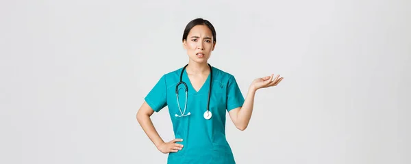 Covid-19, healthcare workers, pandemic concept. Frustrated and annoyed asian female doctor, nurse in scrubs raise one hand and looking questioned and pissed-off, complaining something — стоковое фото