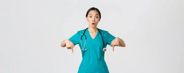 Ковид-19, медицинские работники, пандемия. Удивлённая и впечатлённая азиатская медсестра, доктор в халатах, показывающая пальцами вниз и выглядящая сомнительно, заинтересованная в новом промо, белом фоне — стоковое фото