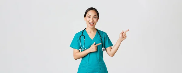 Covid-19, trabajadores de la salud, concepto pandémico. Feliz sonriente, hermosa médico asiático, terapeuta femenina en matorrales o enfermera señalando los dedos esquina superior derecha, mostrando el camino al paciente, fondo blanco — Foto de Stock