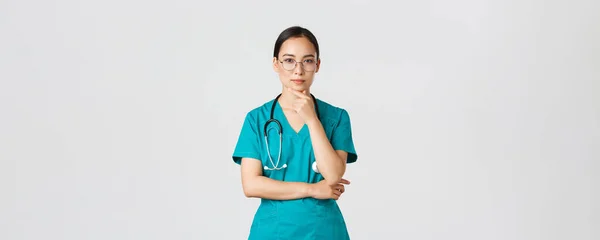 Covid-19, εργαζόμενοι στην υγειονομική περίθαλψη, πανδημία έννοια. Σοβαρή εμφάνιση νεαρή γυναίκα στο διαδίκτυο, ασιατική νοσοκόμα ή γιατρός σε scrubs και γυαλιά σκέψης, μελετώντας ή κάνοντας την επιλογή, εξετάζει τον ασθενή — Φωτογραφία Αρχείου