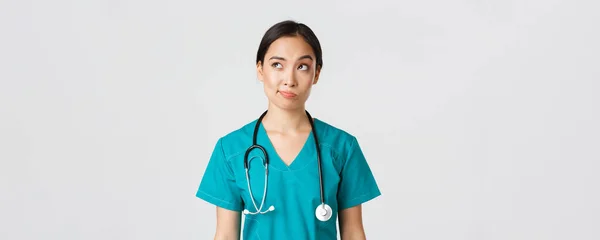 Healthcare workers, preventing virus, quarantine campaign concept. Thoughtful asian female doctor, nurse in scrubs looking upper left corner and smirk unsure, thinking, white background — Stock Photo, Image