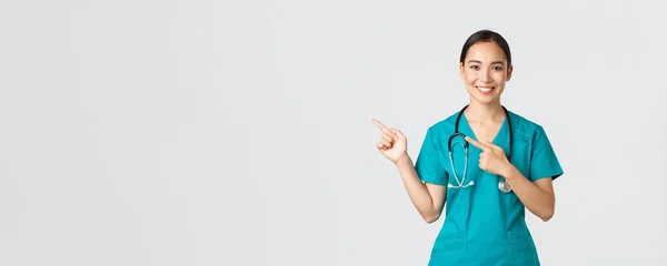 Covid-19, zdravotníci, pandemický koncept. Usmívající se pěkný asijský lékař, zdravotní sestra v drhnutí a stetoskop, ukazující prsty v levém horním rohu, ukazující online klinika banner, bílé pozadí — Stock fotografie